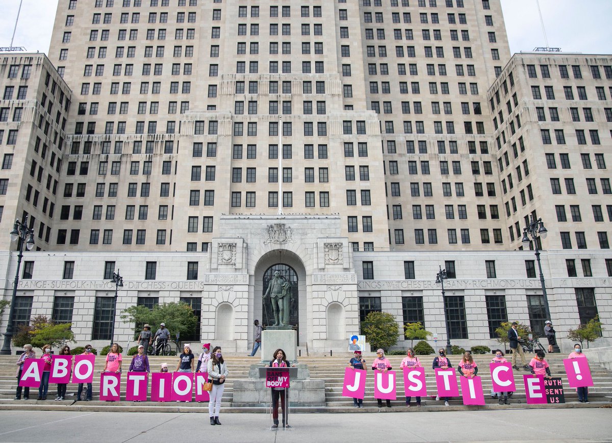 Governor Hochul stands in front of building with people holding signs reading "Abortion Justice!"