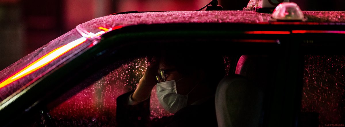 Photography by Liam Wong of Tokyo at night. A taxi driver wearing a mask lit up in red light with his hand resting on his head. He's wearing a face mask. The windows are covered in rain.