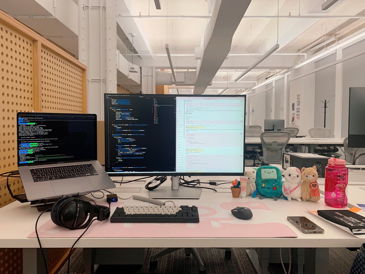 Lauren's desk at work. Pink desk mat, laptop on a stand, 32" 4K monitor. Lots of plushies including BMO and alpacas.