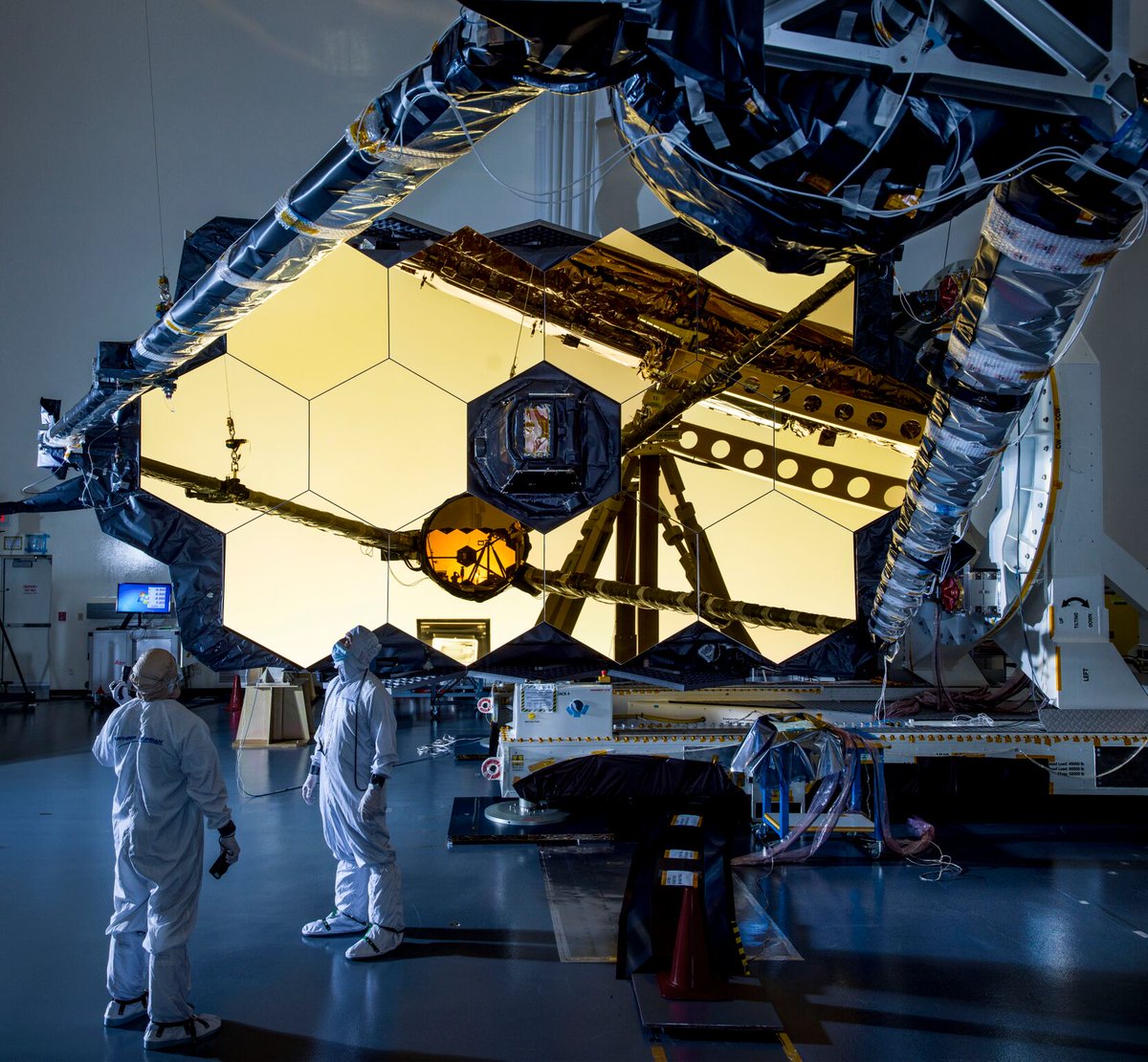 The golden, hexagonal segments of the Webb Telescope's primary mirror have been folded and laid on the ground. The secondary mirror's support structure, resembling arms, has also been extended outwards in front of the primary mirror. Although it does not face the viewer, the small, circular secondary mirror is visible in the reflection of the primary mirror. On the bottom left, technicians and engineers in cleanroom suits visually inspect the support structure of the secondary mirror.