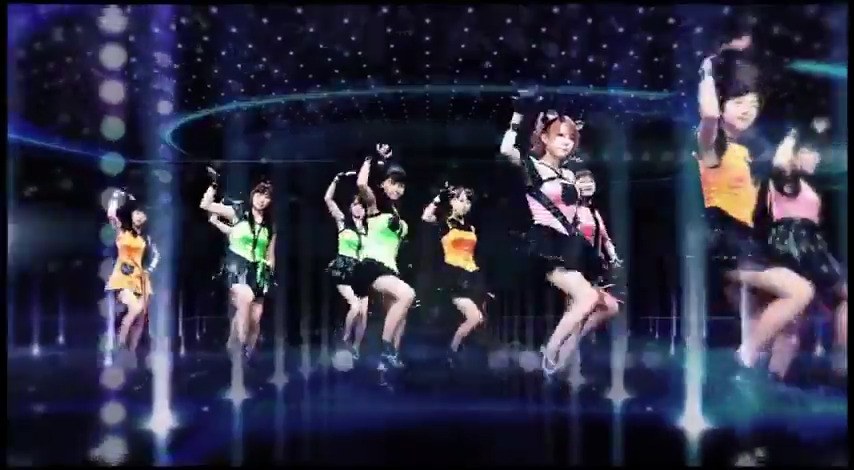 A photograph of a J-pop or idol group performance showing several female performers in colorful outfits (green, pink, orange) dancing on stage with lighting effects and a starry backdrop.