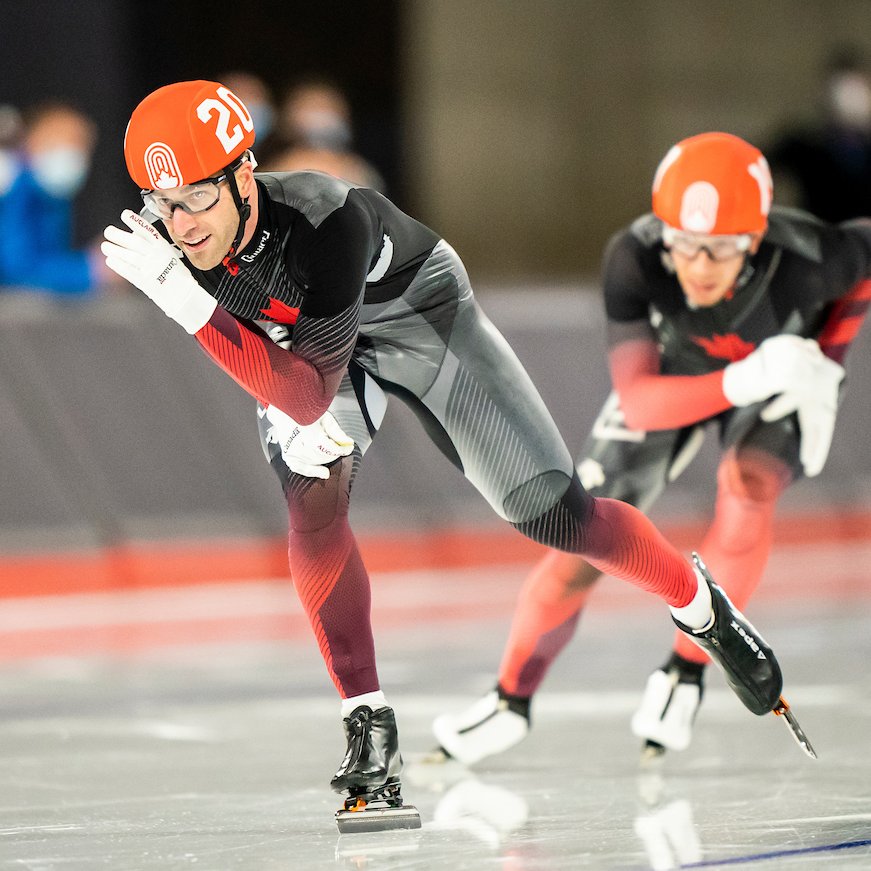 Antoine Gelinas-Beaulieu competing in the mass start event. His right foot is on the ground and his left foot is slightly above the ice. His left arm is in front of his face.