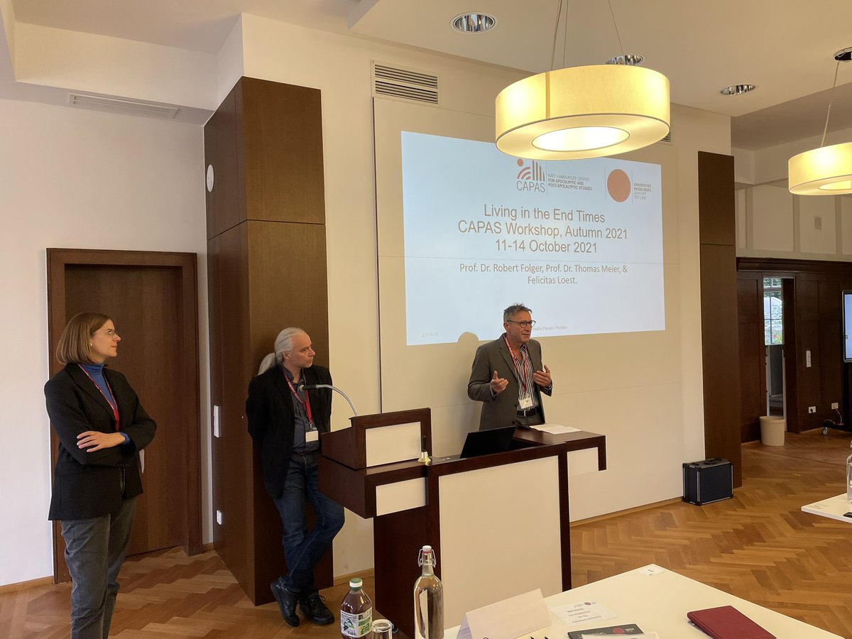 A women and two men are standing in a lecture room with wooden floor at the front. They are talking to an audience not shown in the picture.