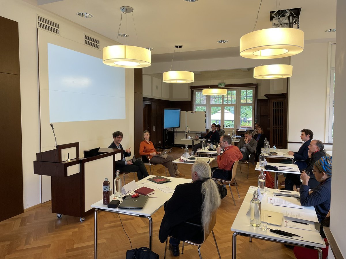 Several men and women are sitting at individual desks in a lecture room. two persons are sitting on chairs at the front discussing with the audience.