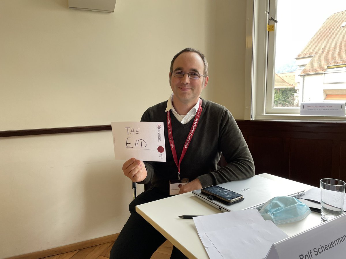 A man is sitting at a table. he is smiling into the camera and holding up a paper, written on it: The End.