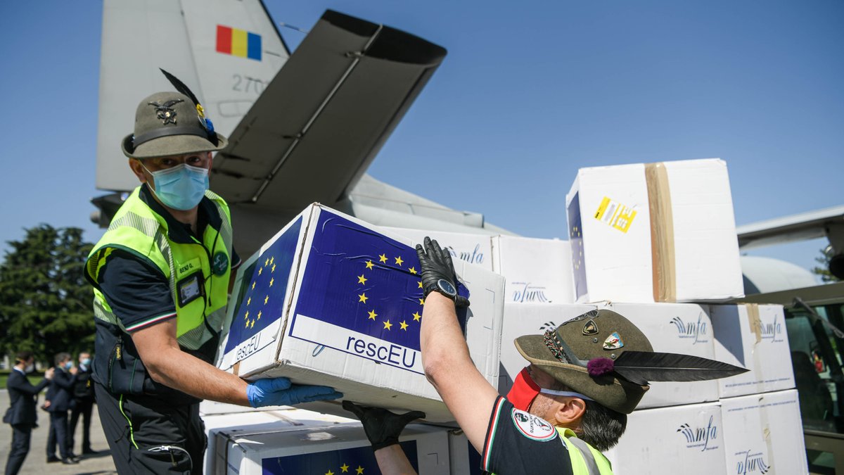 Two people holding a white box with an EU flag stamp on it, where it’s written “rescEU”. These two people wear an alpine hat and are surrounded by other boxes and an airplane.