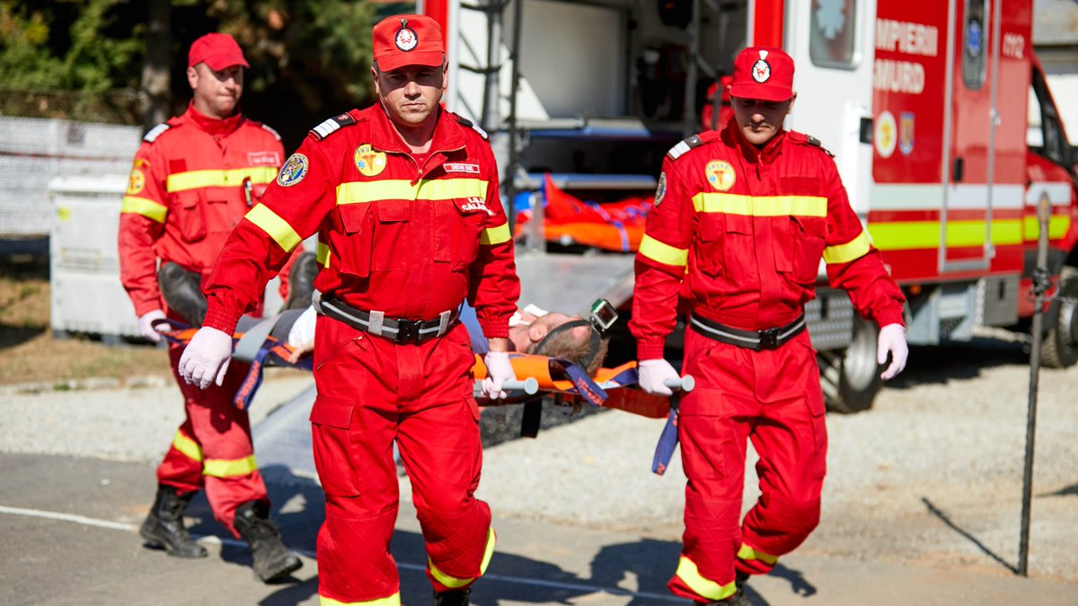 Four people in a red uniform practicing a rescuing exercise. An ambulance can be seen in the background.