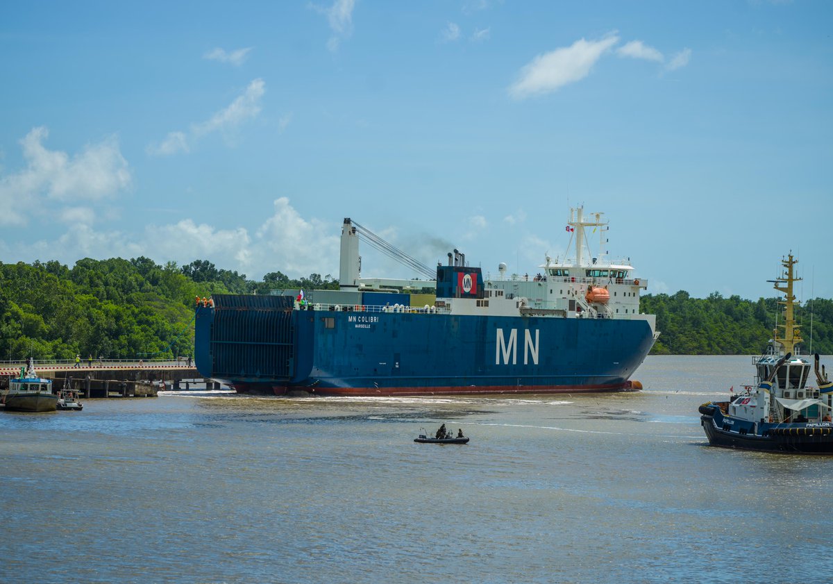 A large, blue cargo ship with the words MN Colibri written on the side sits docked at a port with several shipping containers loaded on the top of it – one holding the James Webb Space Telescope. The sky is blue with clouds and green trees line the coast.  