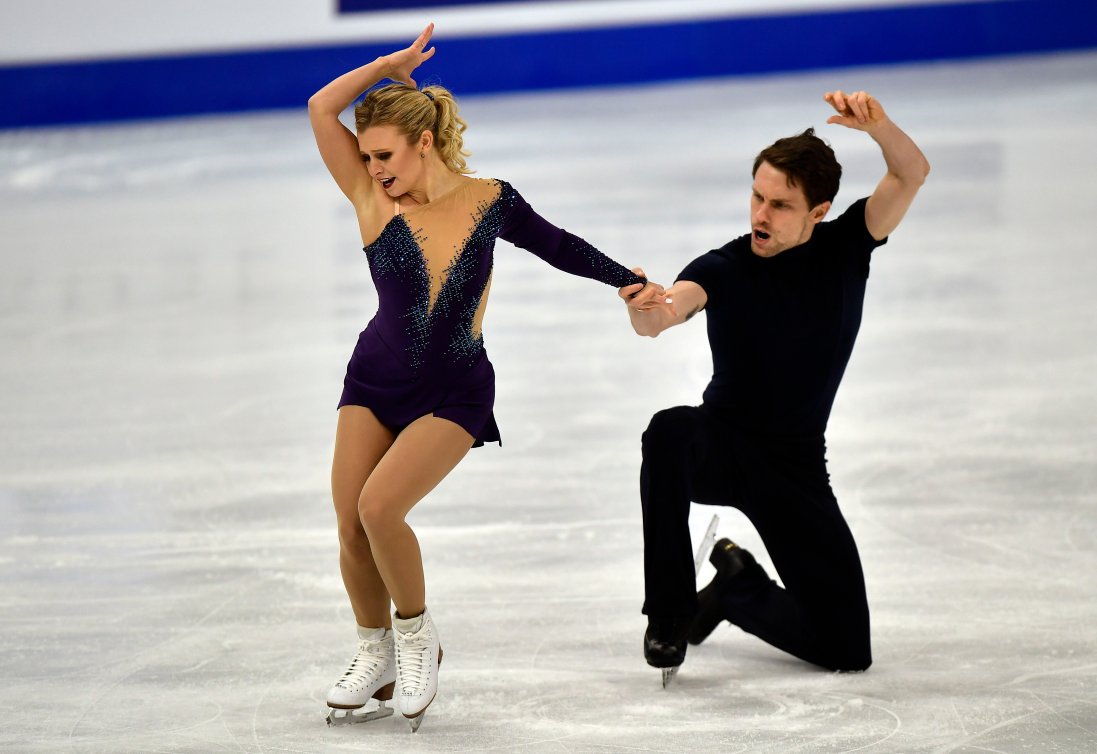 Kirsten Moore-Towers (left) and Michael Marinaro pose on the ice. Kirsten has her right hand in the air and her left knees bent slightly. Michael is holding onto her left hand is is kneeling on the ice behind her.