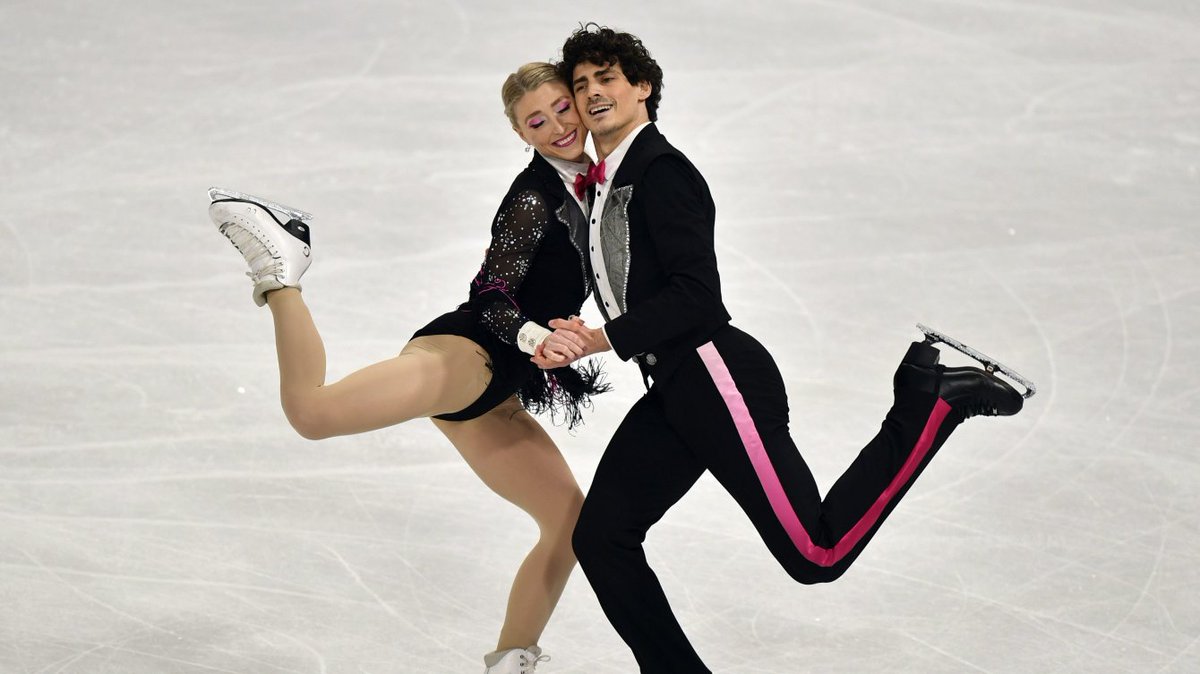 Piper Gilles (left) and Paul Poirier hold hands and lean their bodies towards each other while each has a foot in the air. 