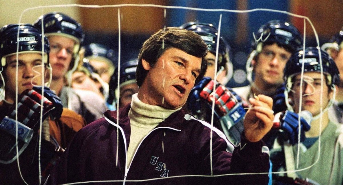A male coach draws on the hockey boards to explain something to his players. The players are crowded around him watching.