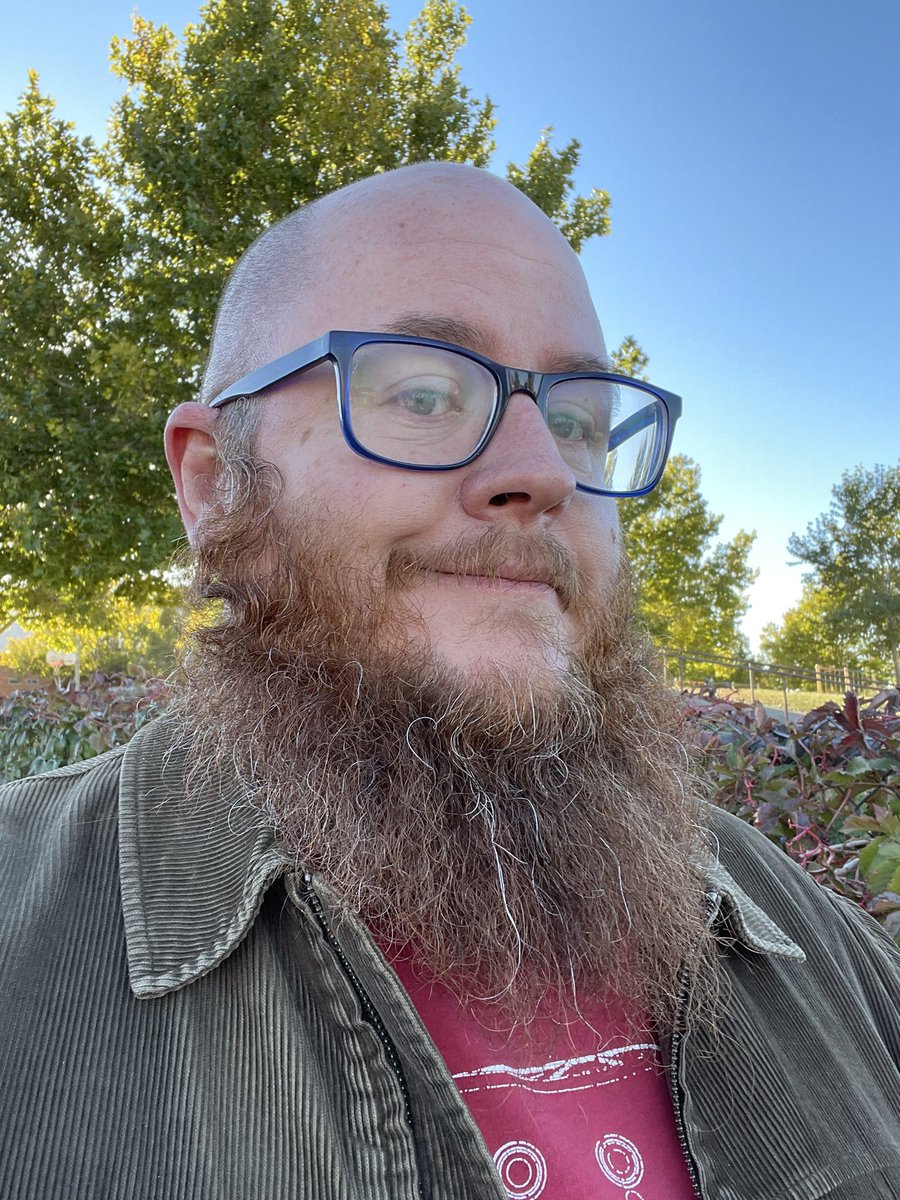 Selfie, red beard all wild with a few streak of gray, corduroy jacket, big smile with vivid green trees and a blue sky behind me