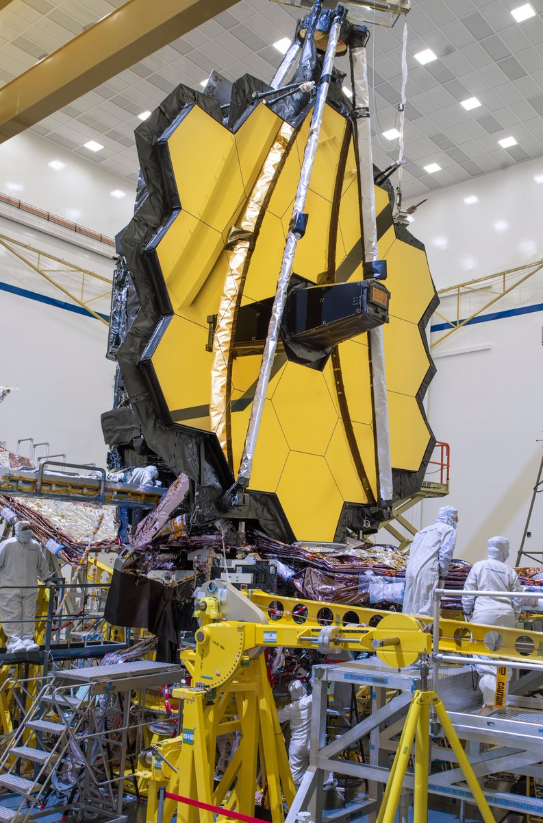 The James Webb Space Telescope with its fully deployed primary mirror. The golden mirror resembles a honeycomb with 18 hexagonal segments. Two technicians in cleanroom suits stand on platforms towards the right.