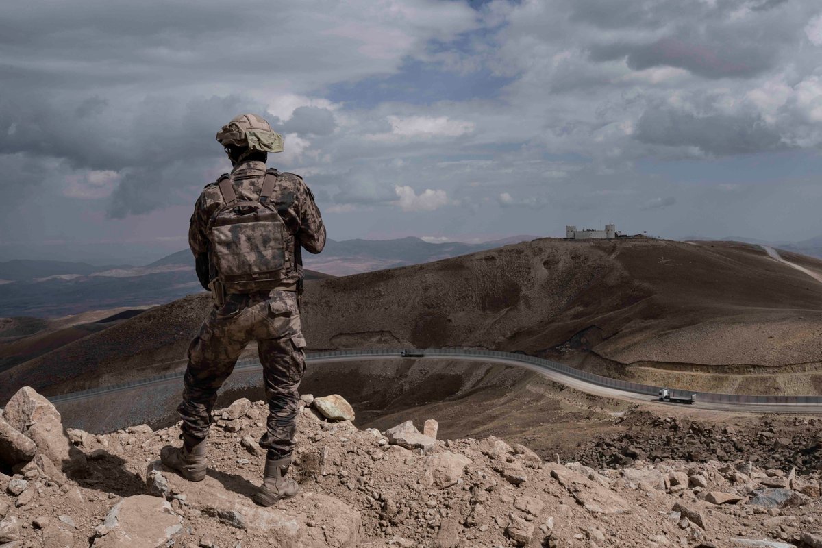 A soldier stands on the top of a hill, looking at the border wall between Turkey and Iran, with his back to the camera