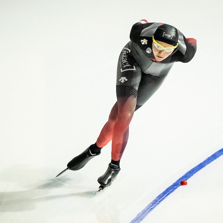 Long track speed skater Isabelle Weidemann skating on the ice during a race. Her hands are behind her back and she is leaning forward, bent forward at the waist.
