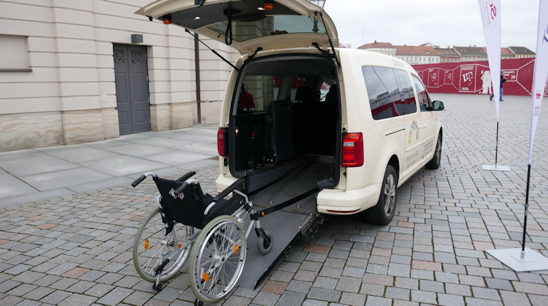 Bildbeschreibung: Ein Taxi mit einer Rollstuhlrampe, auf der Rampe steht ein schwarzer Rollstuhl.