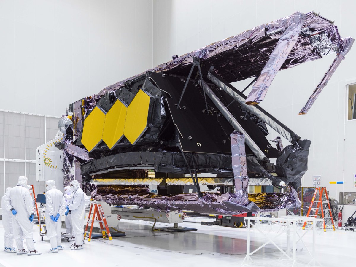 The Webb Telescope laying horizontally in the Guiana Space Center cleanroom after being removed from its container. Technicians stand to the left.