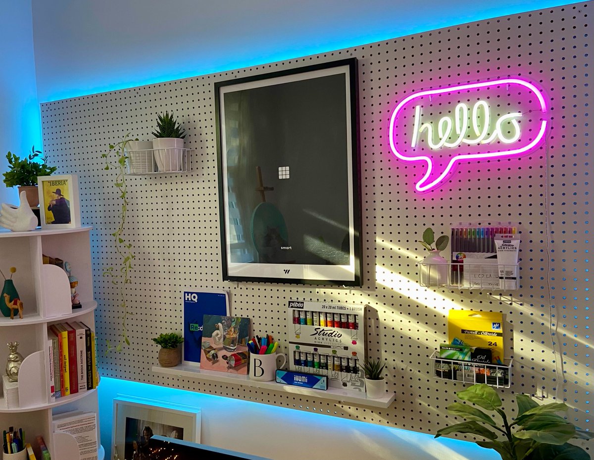 A peg board attached to the wall covered in various Tucows ephemera and art supplies. The main features are a Hello neon sign in yellow cursive test with pink speech bubble, sets of acrylic paint and a Tucows poster that reads "smart"