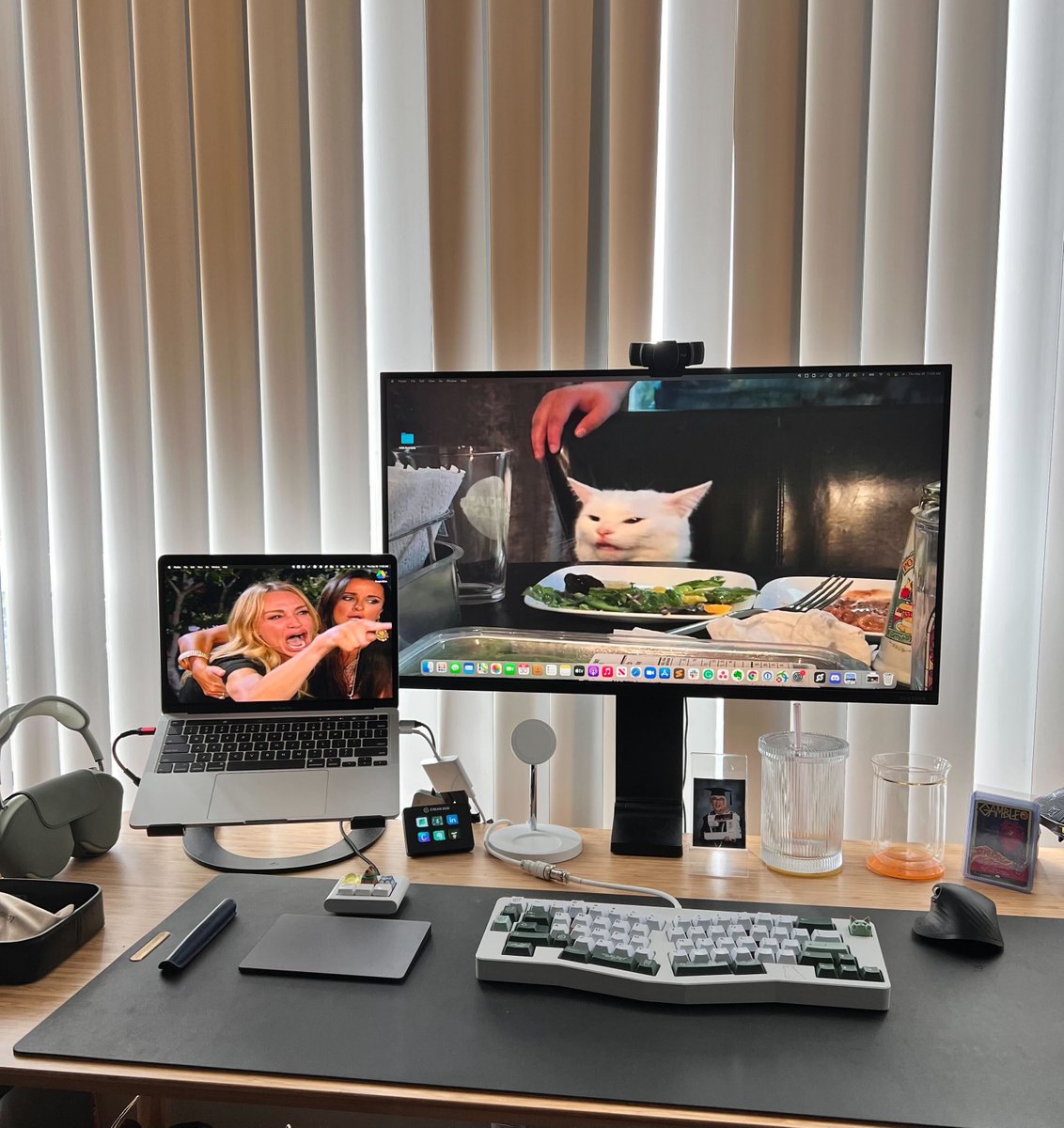  a desk with a macbook pro attached to an external monitor with a logi webcam. There is an ergo keyboard, apple airpods max, Logi Mx3 mouse, apple trackpad and a pen all sitting on a leather desk mat. The screen wallpaper is the ladies yelling at a cat eating a salad.
