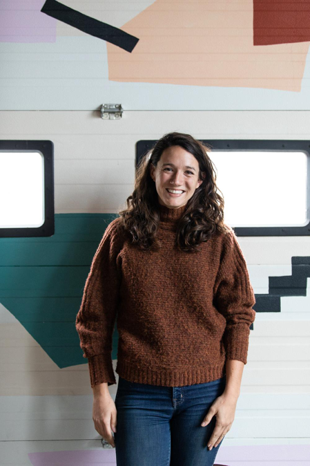 A woman stands in front of a wall, looking to camera. The wall has shapes painted on it in different colours, and two windows