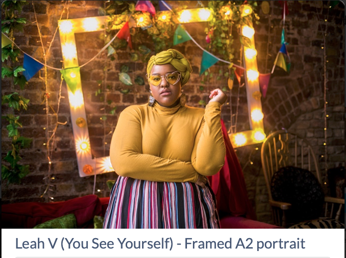 screenshot of a photo with the description "Leah V (You See Yourself) - Framed A2 portrait" below.

The image itself is of a young woman stood centre of frame looking calmly into the camera, one arm across her waist, the other raised at the elbow.  She is stood in front of an exposed brick wall, centred in front of a frame with lit bulbs all round it.