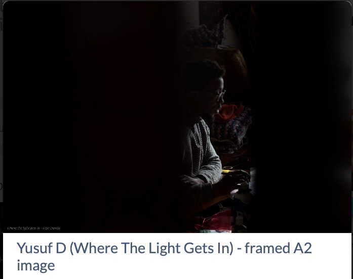 screenshot of an image with the title & description "Yusuf D (Where The Light Gets In) - framed A2 image" underneath.
The photo itself is 3/4 in darkness. in the strip of light right of centre, you can see a young man sat, looking intently at something not seen.