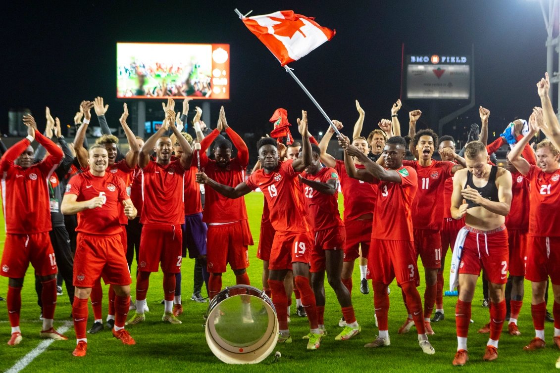 The men's soccer team raises their hands in the air while huddled together. One person is waving a Canadian flag in the air. The sky is dark and they are standing on the field at BMO Field.