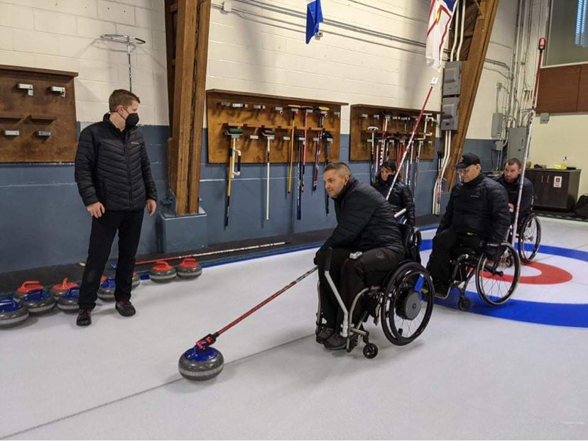 4 adults, 3 male 1 female, in all black clothing seated in wheelchairs on a sheet of curling ice. Each holding push sticks to send their rocks down to the house. An able-bodied, male coach stands to left wearing a mask giving instructions.