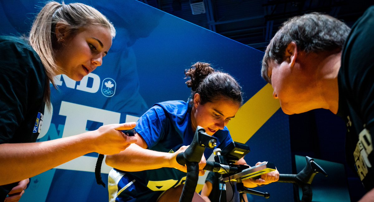 A low angle photo of an athlete riding a bike while a woman (left) holds a stopwatch and a man (right) looks on.