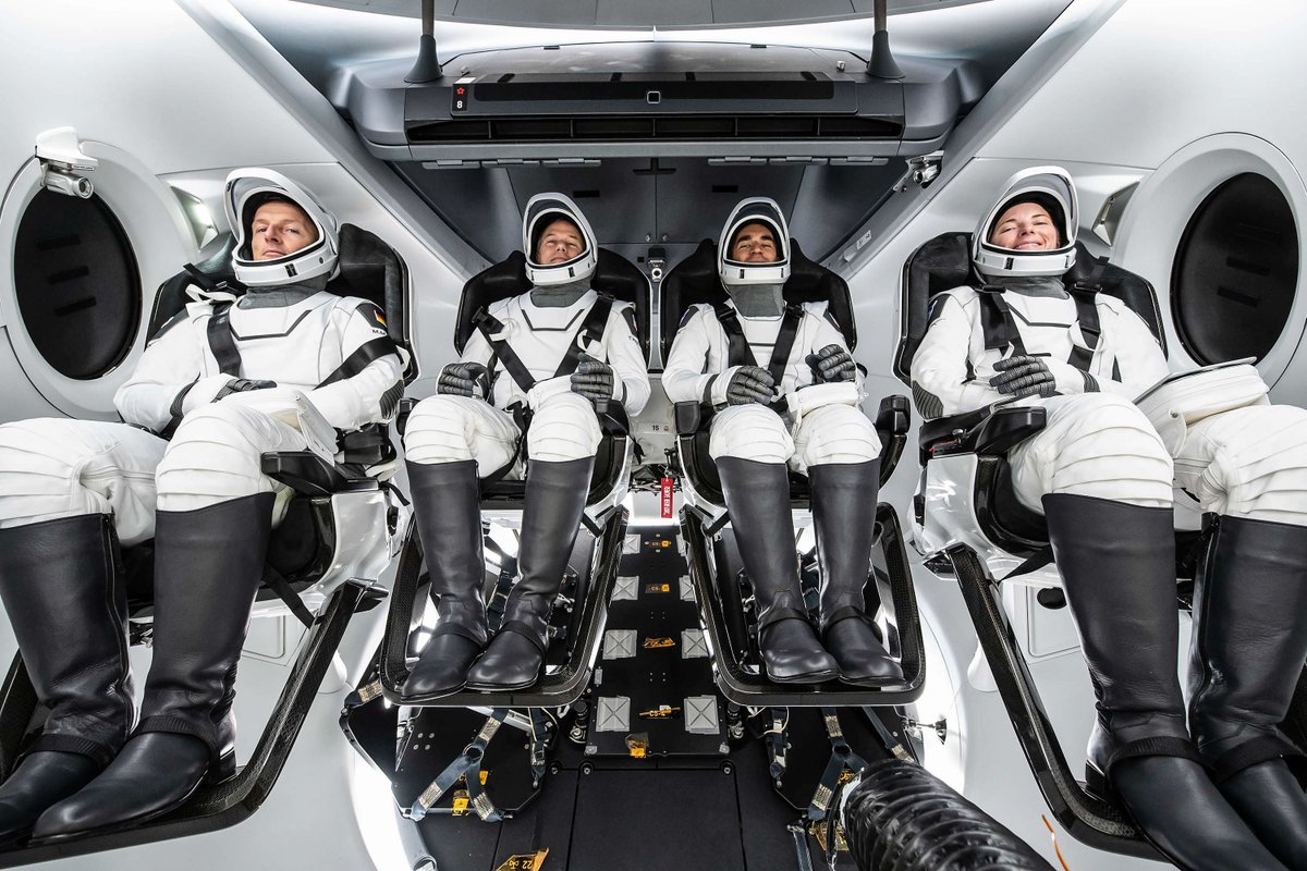 Four astronauts (the crew of NASA's Crew-3 mission), in white launch suits with open helmets and black boots, strapped into SpaceX's Dragon spacecraft, with white walls and round windows on each side