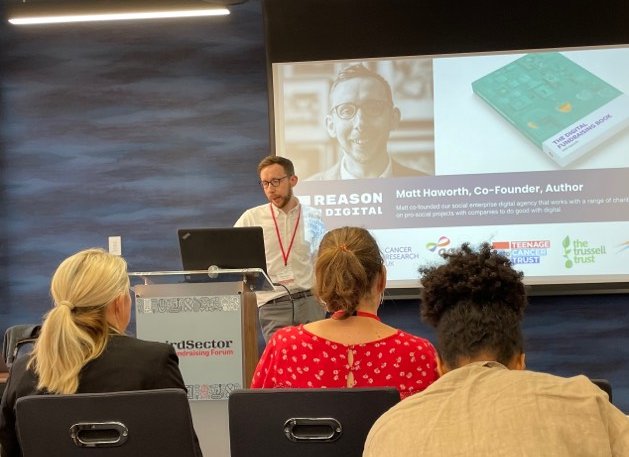 Speaker stood at the front of a room with a screen displaying a presentation at a Third Sector event.