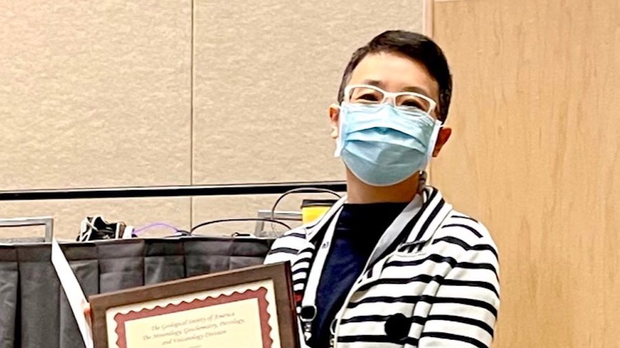 Dr Xiao-Ming Liu looks at the camera and hold up an award. She is standing in a conference room and is wearing a mask.