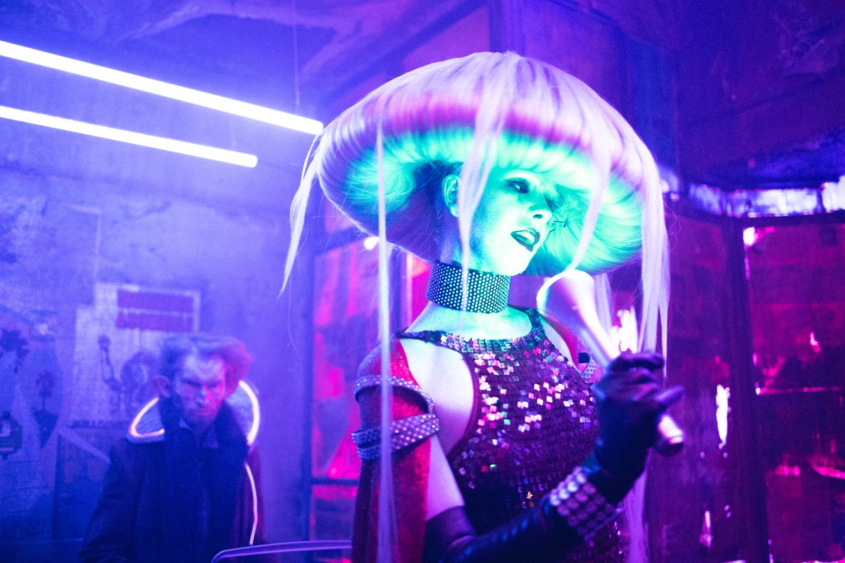 An actor in alien makeup and costume. The actor is wearing a wig that looks like a mushroom top. They are glowing and almost look like a jelly fish. 