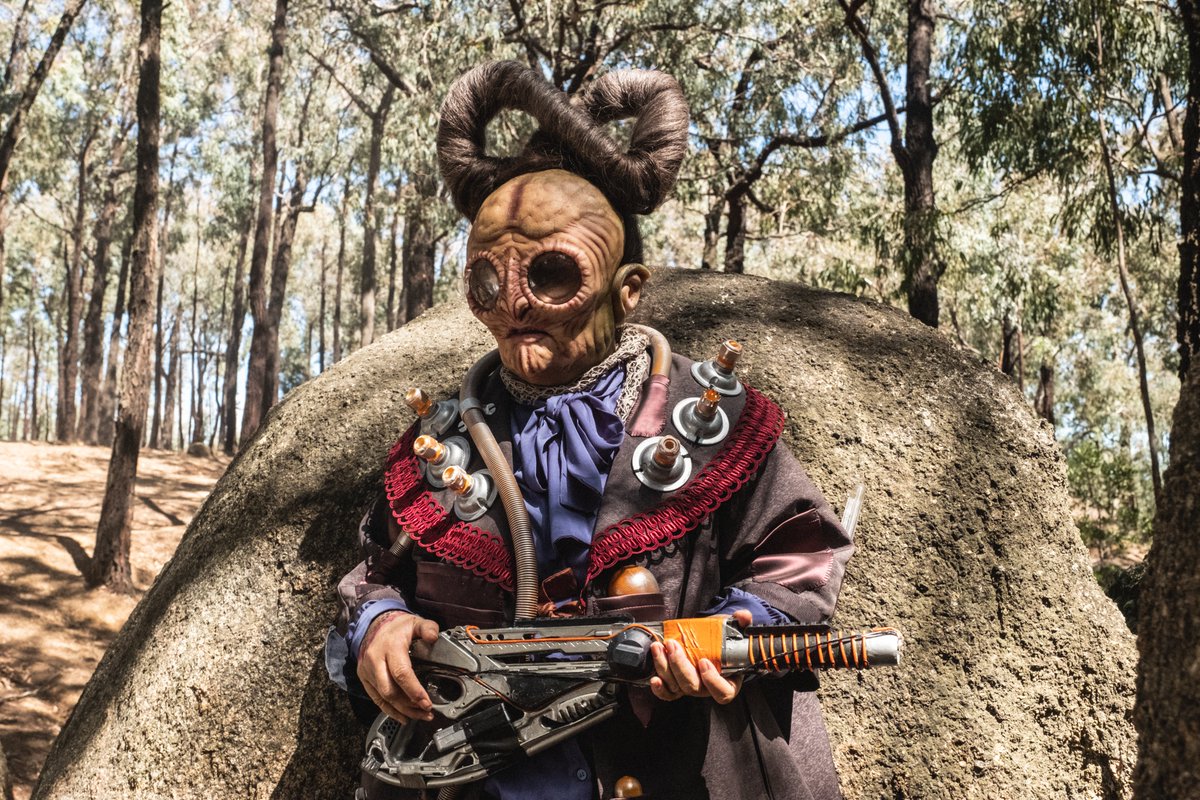 An actor in alien makeup and costume. The actor is wearing a mask that looks like their eyes are giant glass lenses. Their head is bald and looks like their brain is exposed. On top of their head is an infinity sign made out of hair.