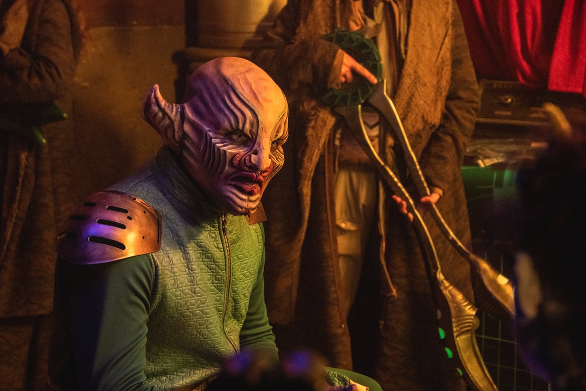 An actor in alien makeup and costume. His alien scalp is bald and veiny. He has horns where his human ears would be. He looks angry.