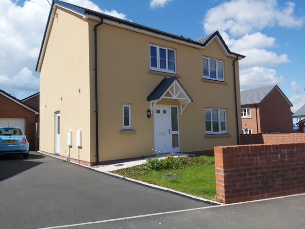 On the left hand side is a drive with a small blue car. Next to that is a house covered in a yellow render. In front of the house is a grassy front garden and a brick wall. 