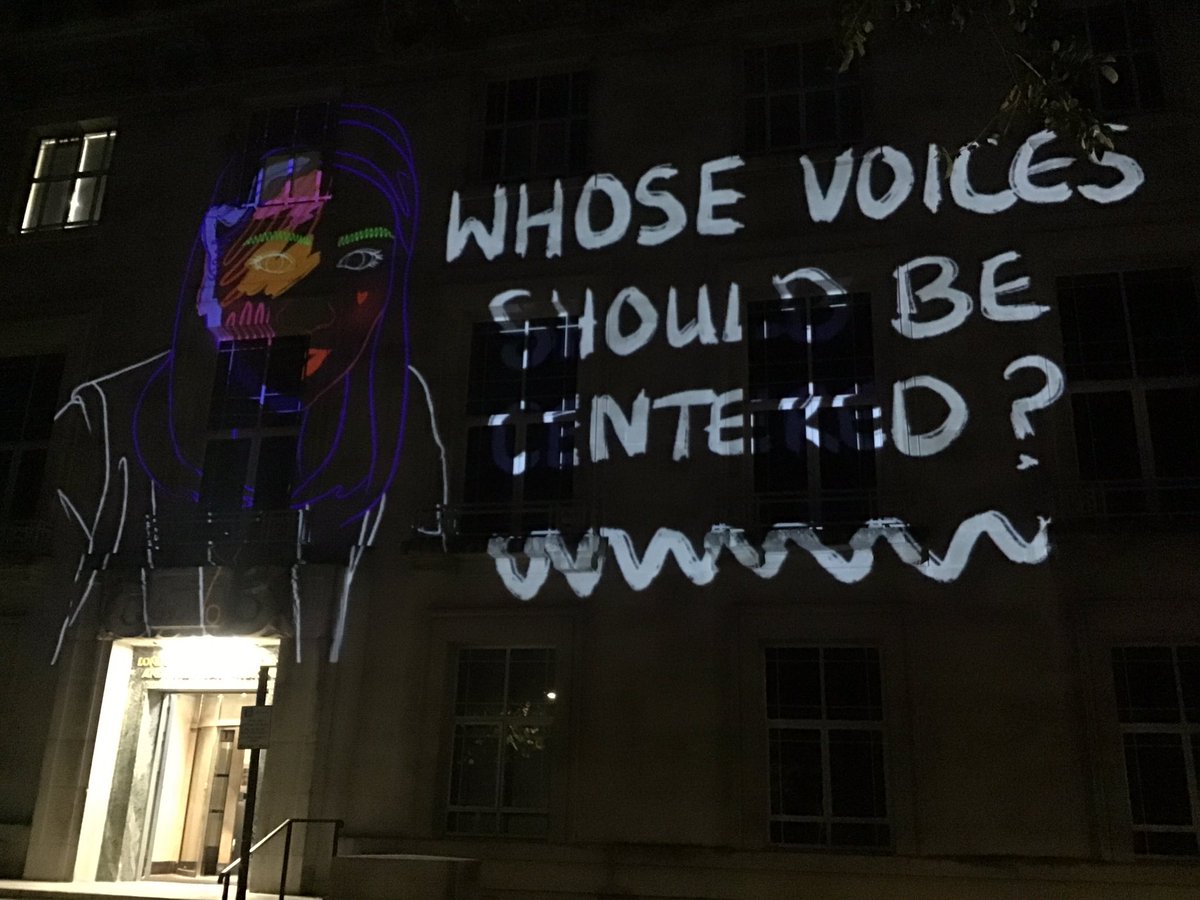 Another of the neon projections - text: “Whose voices should be listened to?”
