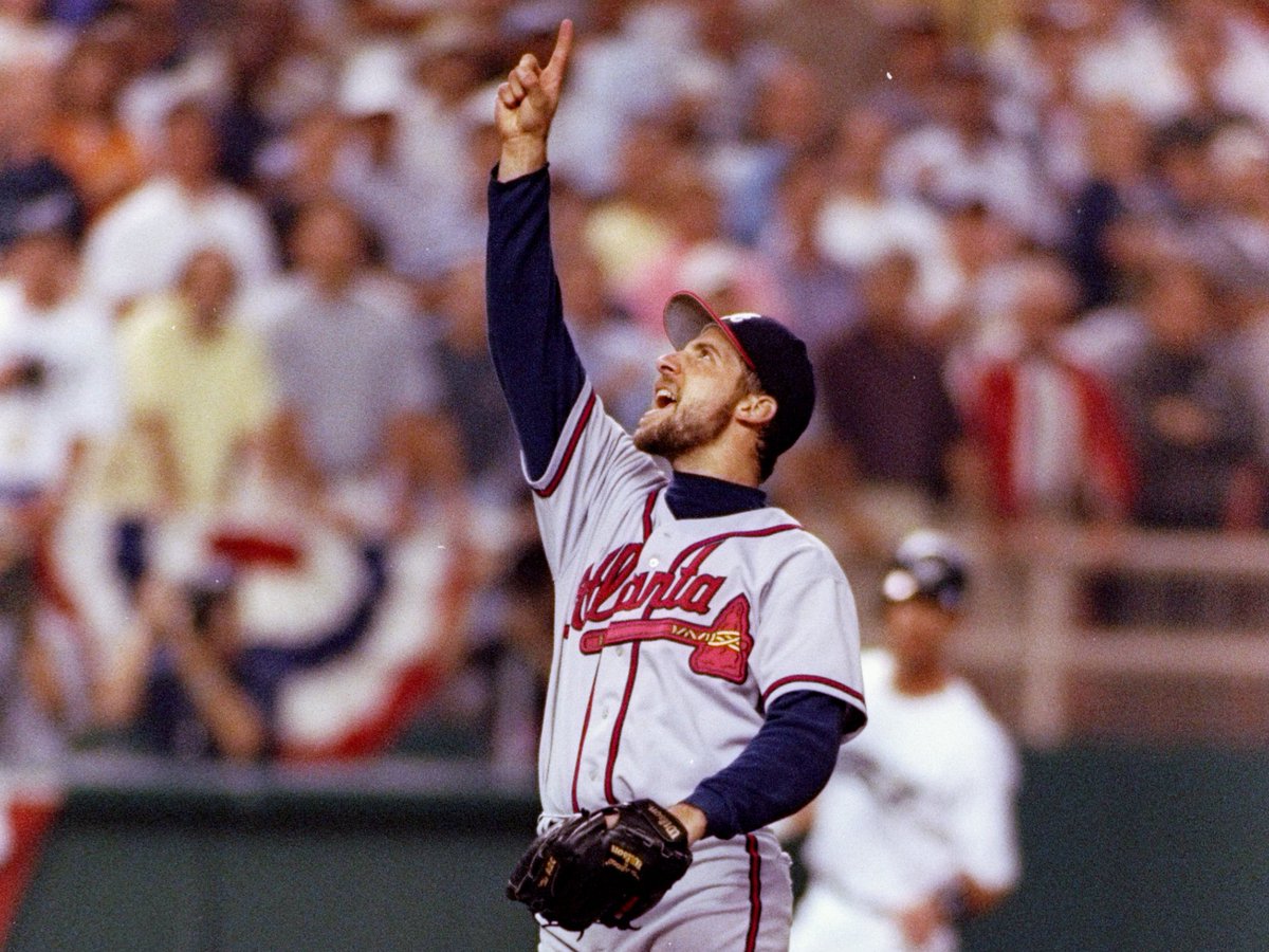 An Atlanta Braves baseball player in a gray away uniform with the team's tomahawk logo pointing skyward in celebration, wearing a baseball glove with a blurred crowd visible in the background.