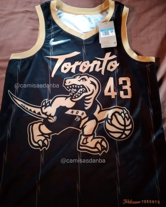 Camisa City Edition preta do Toronto Raptors vista de frente. Ela tem gola e mangas douradas, o logotipo brilhante da Nike na altura do ombro direito. No peito, estão o logotipo Toronto, o número 43 e o desenho de um dinossauro quicando uma bola de basquete. Todos esses detalhe na cor dourado. A camisa tem listras finas em tom cinza, que fazem uma forma similar a um zigue-zague. A camisa está sobre uma cama de cor marrom. A imagem tem marcas d'água do perfil Camisas da NBA.