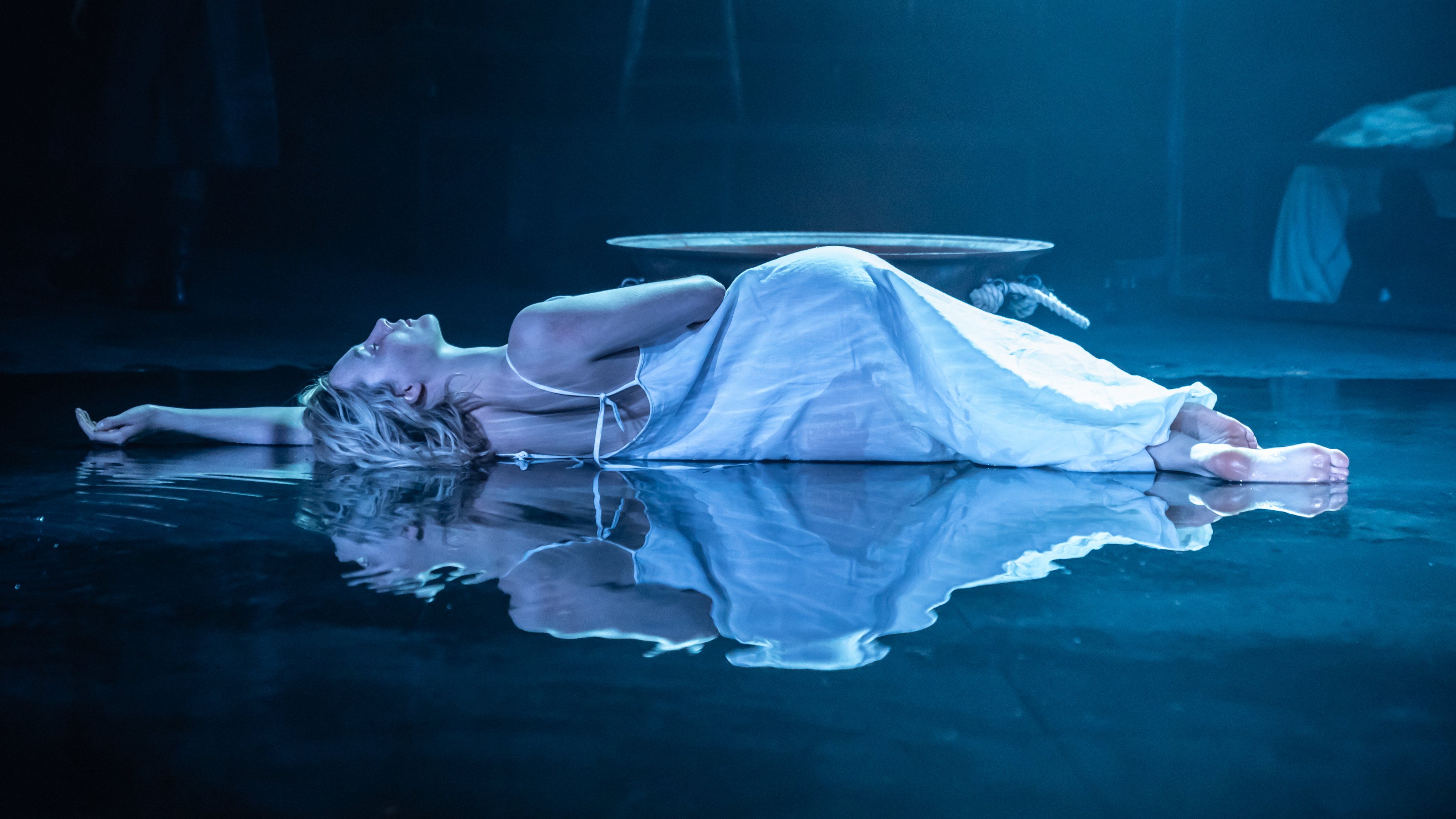 Lady Macbeth (Saoirse Ronan) lays down on a flooded stage. She wears a white dress and has her eyes closed. We see her reflection in the water.