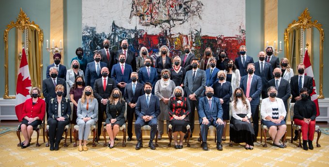 Forty people are split into four rows for a group photo in the Ballroom of Rideau Hall. The Prime Minister and the Governor General are in the middle of the front row. There is a large painting on the wall behind the group, and a Canadian flag on either side.