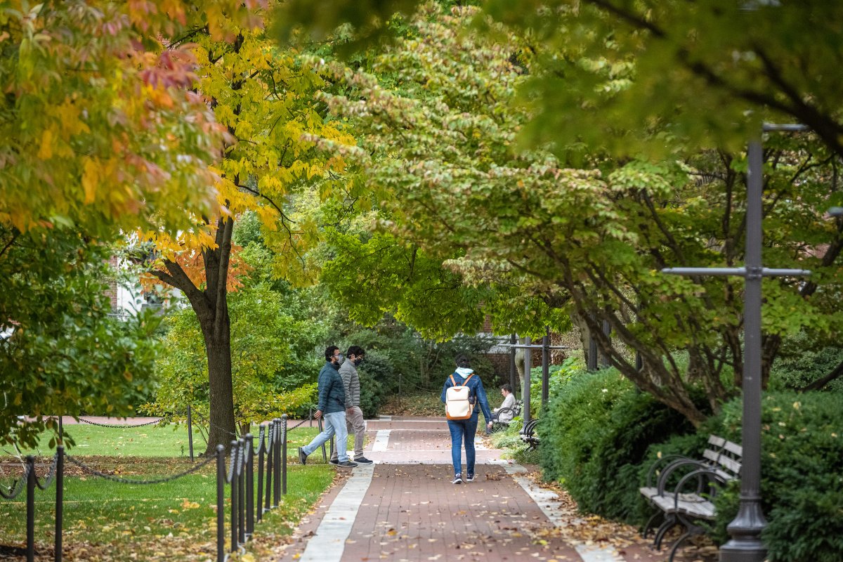 Students walk across the Johns Hopkins University campus. Trees with autumn leaves line the brick pathway.