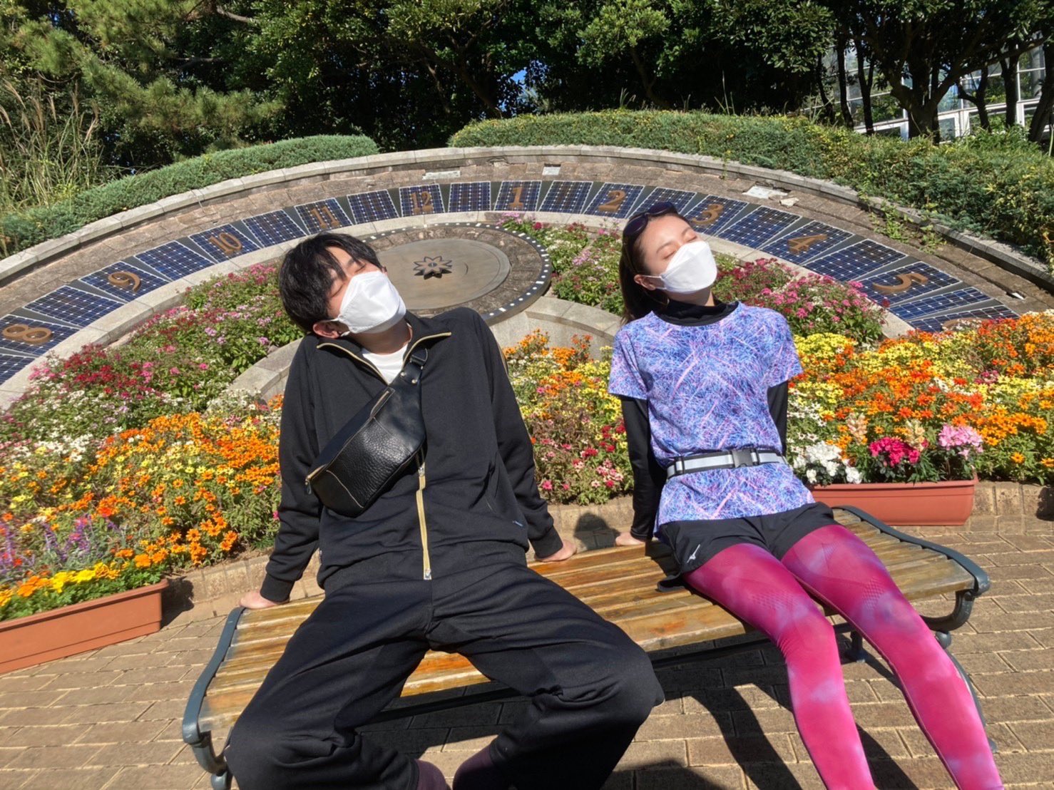 6話のオフショット写真。場所は花がたくさん咲いている公園。モリオ役のスギノヨウスケさんとソラ役のタナベモモコさんが並んでベンチに座っている。左側にスギノさん、右側にタナベさん。2人ともベンチの後ろに両手を置いてのけぞるような体勢で、首を少しかしげながら、目をつむっている。スギノさんは黒のジャージ上下に、黒のボディバッグを身につけている。田辺さんは、黒の長袖インナーに紫のシャツを重ね着して、短パンにピンクのスパッツを履いている。2人とも白いマスクをしている。