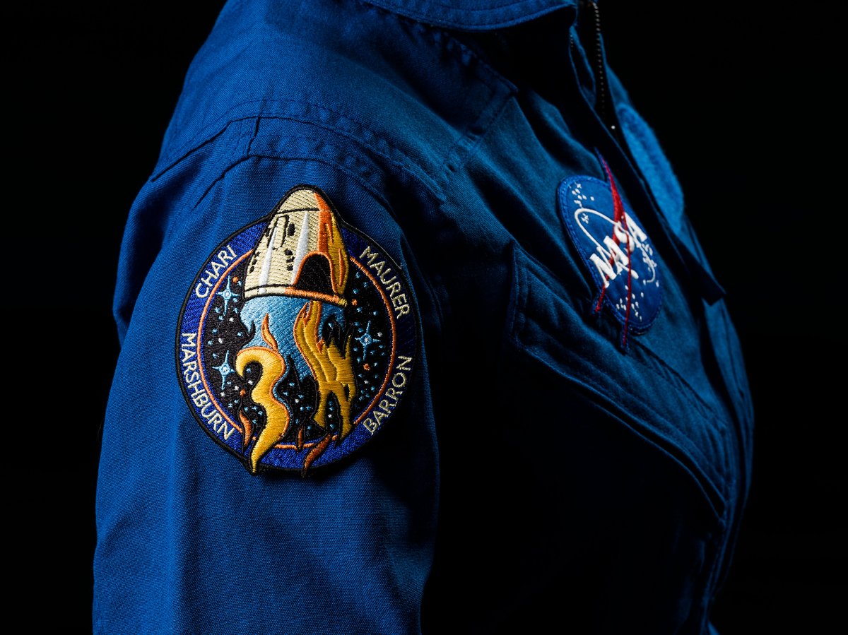 Photograph of blue NASA astronaut flight suit from the side with SpaceX Crew-3 mission patch on upper arm. Patch design includes Crew Dragon spacecraft, astronaut names, stars and planets and a large number 3.