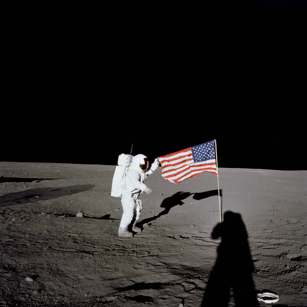 An astronaut holds the end of an American flag on the grey lunar surface. 