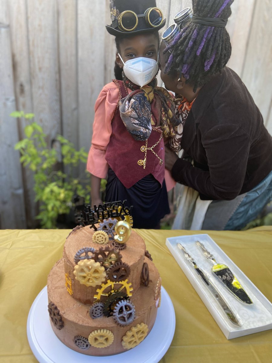 Black girl in steampunk costume with her mother whispering in her ear. They are behind a table with a 2-tier steampunk-themed chocolate birthday cake.