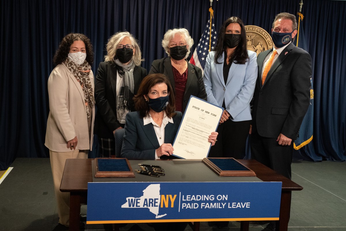Governor Hochul sits while holding signed legislation with five people standing behind her.