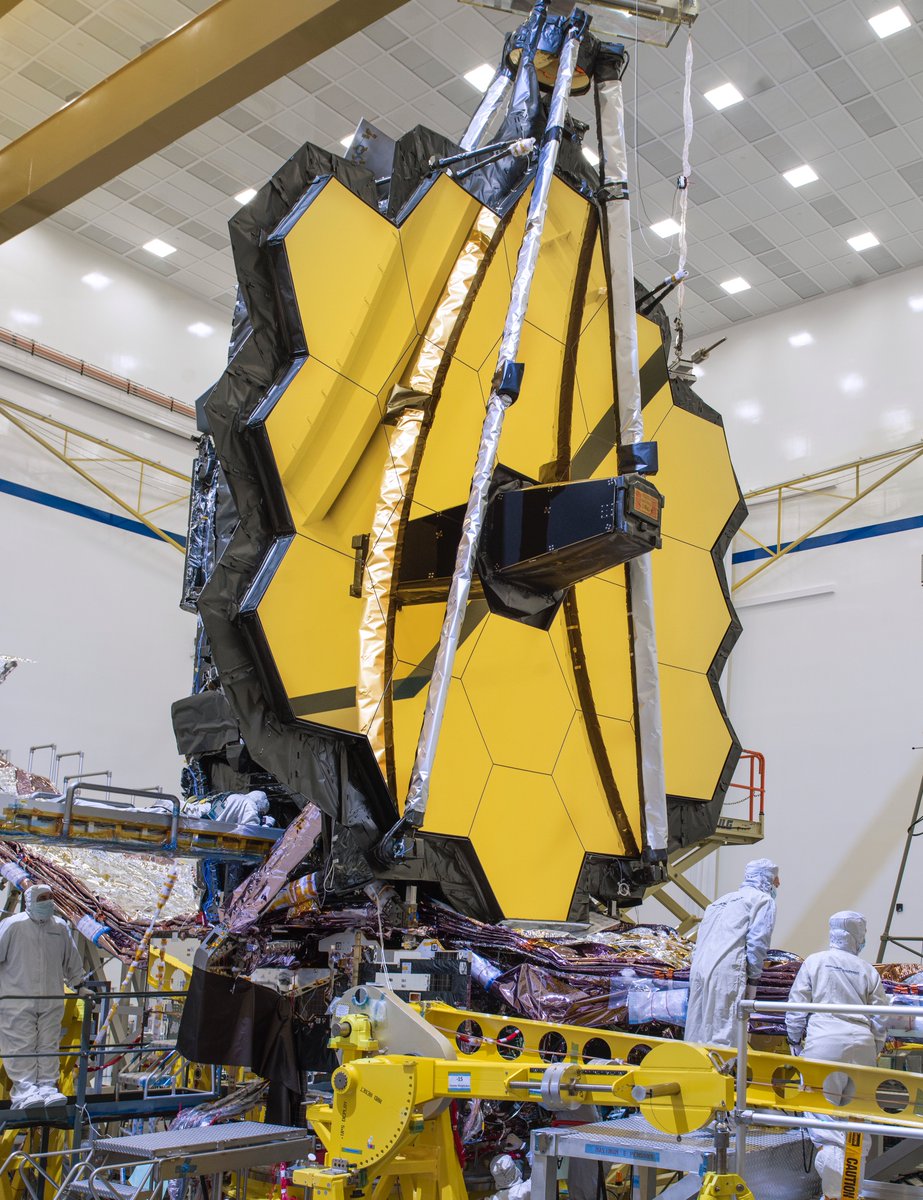 The James Webb Space Telescope with its fully deployed primary mirror, which resembles 18 golden hexagonal segments pieced together into one large honeycomb. Several technicians stand around the telescope.