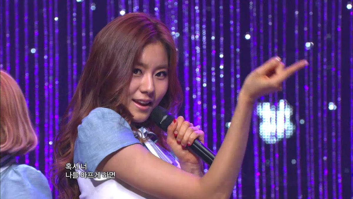 A young woman with reddish-brown hair performs on stage, holding a microphone and pointing, with Korean subtitles '혹시 너 나를 아프게 하면' visible on screen against a sparkly purple curtain backdrop.