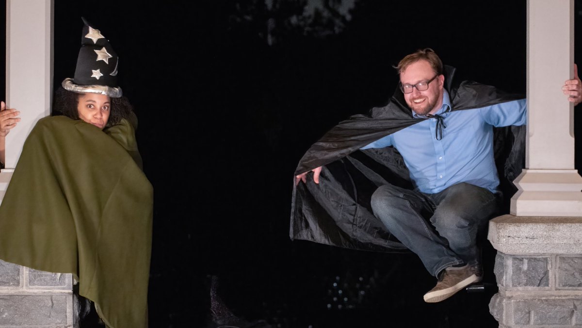 Moiya wears a green cloak and a blue wizard’s hat with silver stars and moons. Rob wears a black cape. Each clings to a pillar, Moiya on the left, and Rob on the right.